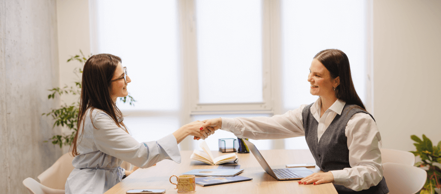 Two businesswomen shaking hands across a table in a modern office setting, symbolizing partnership and collaboration.