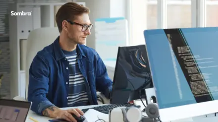 A male working at a desk in an open-space office setting, looking at a computer monitor